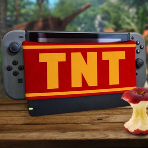 Puede incluir: Una consola Nintendo Switch negra con una funda de tela roja y amarilla que dice "TNT". Una manzana roja a medio comer está en primer plano.