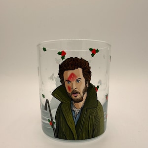 Könnte beinhalten: Ein klares Glas mit einer Cartoon-Illustration eines Mannes mit einem roten Fleck auf der Stirn und einem grünen Mantel. Das Glas ist mit Stechpalmen und einem Brecheisen verziert. Das Design ist vor einem schneebedeckten Hintergrund.