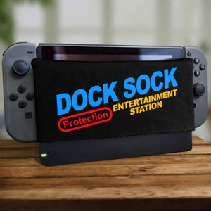 Puede incluir: Un protector de base negro para una consola Nintendo Switch. El protector tiene el texto "Dock Sock" en letras azules y amarillas, y el texto "Protection Entertainment Station" en letras amarillas.