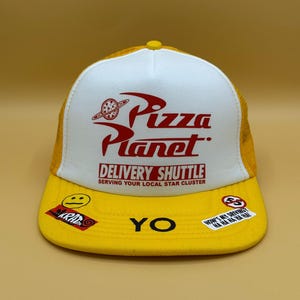 Peut inclure: Une casquette trucker jaune et blanche arborant un logo rouge Pizza Planet avec le texte DELIVERY SHUTTLE SERVING YOUR LOCAL STAR CLUSTER. La visière jaune affiche le mot YO et plusieurs petits autocollants dont un smiley et du texte.