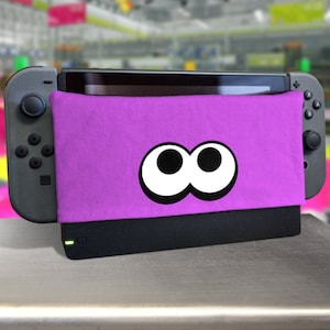 Könnte beinhalten: Eine violette Nintendo Switch-Konsole mit einem pinkfarbenen und weißen Augen-Design auf dem Etui.