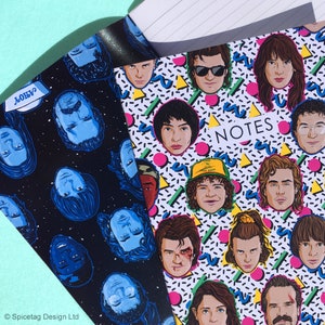 Puede incluir: Dos cuadernos con temática de Stranger Things. Un cuaderno tiene un fondo negro con caras de personajes azules y blancas. El otro cuaderno tiene un fondo blanco con un patrón de estilo de los 90 y caras de personajes coloridos. El texto "NOTES" está impreso en el cuaderno blanco.