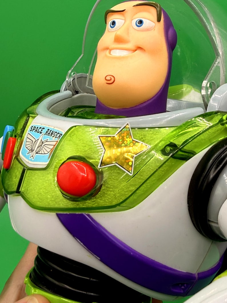 May include: A close-up of a Buzz Lightyear action figure. The figure has a white and green spacesuit with a red button, a gold star, and the words "SPACE RANGER". The figure's head is a light peach color with a purple helmet and a smiling expression.