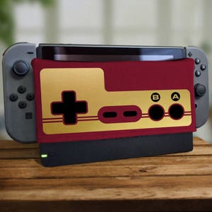 Puede incluir: Una funda para consola Nintendo Switch de color rojo y dorado con un diseño retro de mando de videojuego. La funda presenta un joystick clásico, botones etiquetados con "A" y "B", y un botón con signo más.