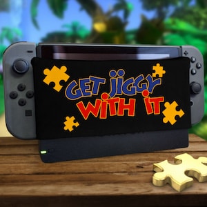 Puede incluir: Una consola Nintendo Switch negra con una piel personalizada que muestra el texto "Get Jiggy With It" en letras azules, rojas y amarillas. La piel también presenta diseños de piezas de rompecabezas amarillas.