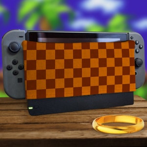 May include: A Nintendo Switch console with a brown and orange checkered cover, a gold ring, and a black charging dock. The background features a blurred green and blue landscape.