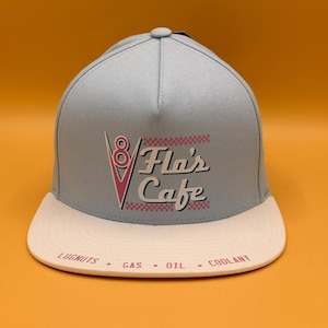 May include: A light blue baseball cap with a flat brim. The front features a graphic with a red V8 logo and the text Flo's Cafe. The brim is off-white and printed with the words LUGNUTS, GAS, OIL, and COOLANT in a red font across the front edge.