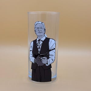 Puede incluir: Un vaso cilíndrico transparente con una ilustración gráfica en blanco y negro de una persona de pelo corto que viste una camisa de cuello, una corbata estampada y un chaleco oscuro, sosteniendo un subfusil con cargador de tambor.