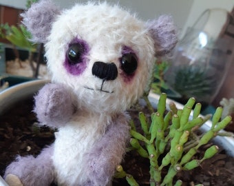 May include: A small, white and purple teddy bear with black eyes and a black nose. The bear is sitting in a silver pot with green plants.