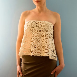 May include: A white crochet top with a delicate, lacy pattern. The top is strapless and has a slightly asymmetrical hemline. The model is wearing a brown skirt.