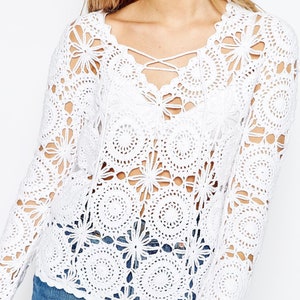 Puede incluir: Top blanco de crochet con escote de encaje y mangas largas. La parte superior está hecha de una tela delicada de tejido abierto y presenta un patrón circular.