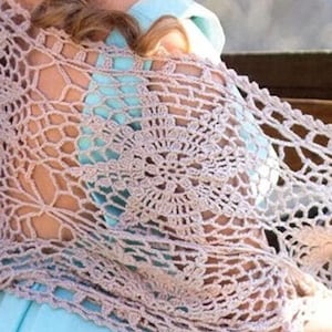 May include: A light brown, open-weave crochet shawl draped over the shoulders. The shawl features a floral pattern and is worn over a light blue dress. The image shows a woman with blonde hair in a bun.