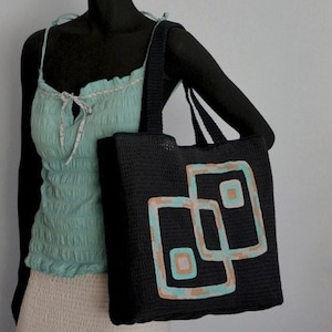 May include: A black crocheted tote bag with a geometric design in teal, peach, and white. The bag has two handles and is shown on a mannequin and on a tripod.