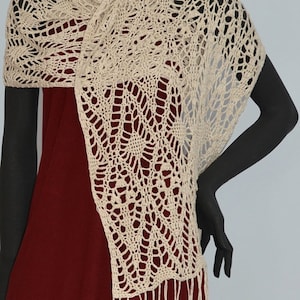 May include: A beige crocheted shawl with a delicate floral pattern and fringe. The shawl is draped over a mannequin wearing a burgundy dress.