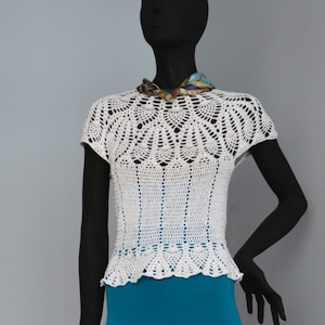 May include: White crocheted top with a scalloped hem and short sleeves. The top is on a mannequin with a teal skirt.