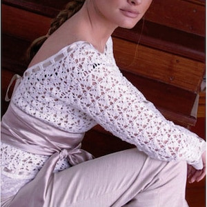 May include: A white crochet top with long sleeves and an open back. A satin sash is tied around the waist. The model is wearing light beige pants. The top features a delicate, openwork pattern.