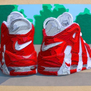 May include: A pair of red and white sneakers made of yarn and nails. The sneakers are sitting on a brown surface with a green background.