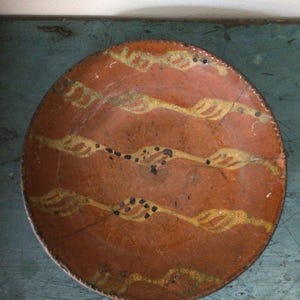 May include: A round, terracotta-colored plate with a decorative design. Three horizontal bands of yellow and white, leaf-like shapes are painted across the plate. Small black dots are interspersed between the bands. The plate has a dark brown rim.