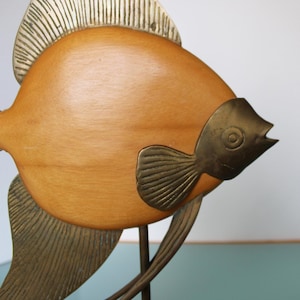 May include: A decorative fish sculpture featuring a smooth, light brown wooden body contrasted with textured brass fins, a brass tail, and a brass head section. The fish is mounted on a thin metal rod, showcasing a combination of wood and metal.