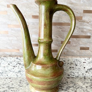 May include: A green and brown ceramic pitcher with a long spout and a handle. The pitcher has a textured surface and a rounded base.