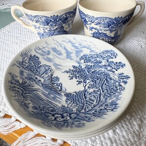 May include: Two cream-colored ceramic teacups and one matching saucer featuring a blue transferware pattern. The design depicts a detailed landscape with trees, a building, and a cloudy sky. The ceramic surface shows a fine crackle glaze finish.