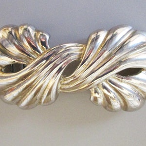 May include: A silver-toned metal belt buckle with a swirling, abstract design. The buckle is shaped like a figure eight and has a smooth, polished finish.