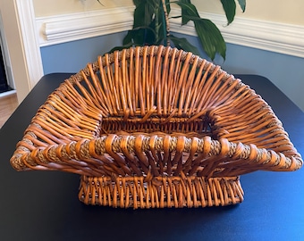 May include: A woven wicker basket with a rectangular shape and a brown color. The basket has a unique design with a raised rim and a flat base.