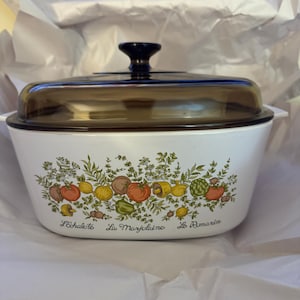 Peut inclure: Plat à gratin blanc avec un couvercle en verre brun. Le plat est décoré d'un motif floral avec des légumes et les mots "L'Echalote", "La Marjolaine" et "Le Romarin".