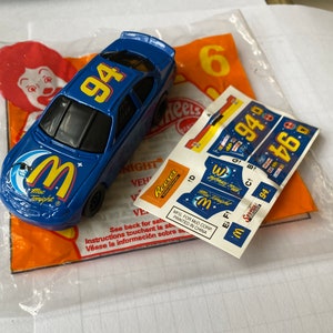 May include: A blue toy race car with a large "M" logo and the words "Mac Tonight" on the side. The car has the number "94" on the roof and a sticker sheet with additional logos and numbers.