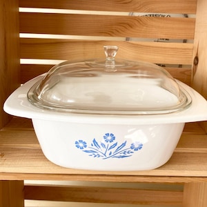May include: White casserole dish with a blue floral design and a clear glass lid. The dish is sitting on a wooden shelf.