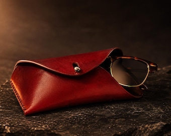 May include: A deep red leather glasses case with a single silver-toned metal snap button closure. The case has a curved flap design and contains a pair of brown tortoiseshell-patterned eyeglasses with thin gold-toned metal frames visible inside.