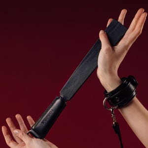 Puede incluir: Paleta de cuero negro con un mango y la palabra "tesstick" en relieve. También se ve un brazalete de cuero negro con un anillo de metal y una correa. El fondo es de color rojo oscuro.