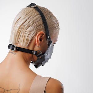 May include: A person wearing a black leather face mask with straps that go around the head and neck. The mask has a metal ring on the side. The person has short blonde hair and a tattoo on their neck that says "balance".