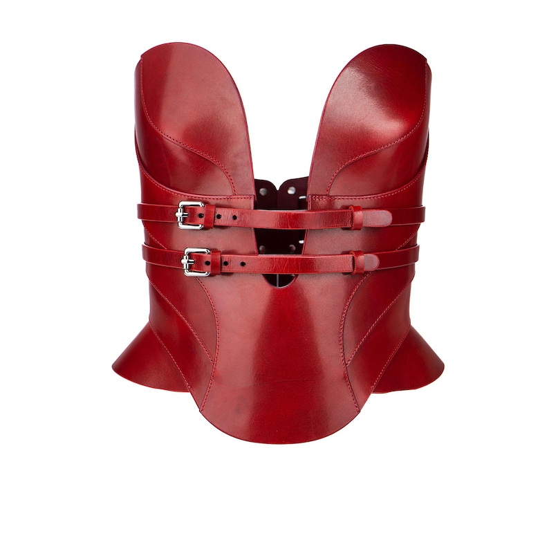 May include: A deep red structured corset top featuring a smooth, glossy finish with two horizontal straps across the front. Each strap is secured with a silver-toned metal buckle. The garment has a high, split neckline and a flared peplum hem.