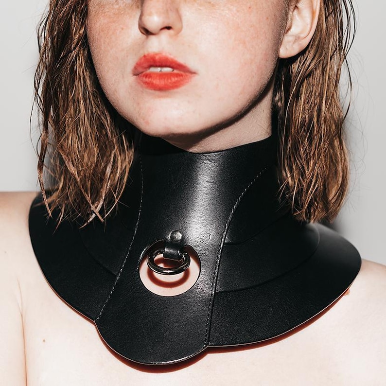 May include: A wide black leather choker collar featuring a layered, curved design that extends over the collarbone. The center of the piece includes a circular cutout with a fixed metal O-ring attached by a small leather loop at the top.