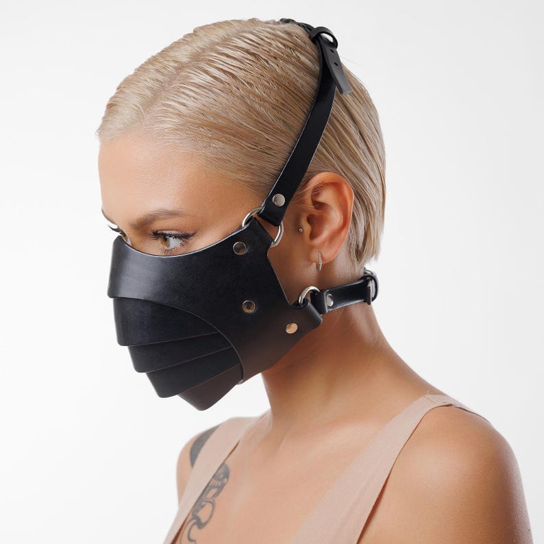 May include: A black leather face mask with a geometric design and adjustable straps. The mask has a layered look and is secured with metal hardware.