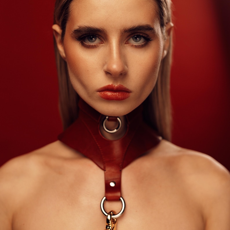 May include: A dark red leather choker collar featuring a wide, contoured neck band with a central silver-toned metal O-ring. A vertical leather strap extends downward from the collar, secured by a silver rivet and ending in a second metal O-ring.