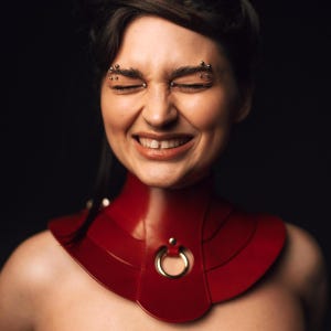 May include: A deep red, structured collar necklace featuring a layered, curved design that sits high on the neck. The piece is accented with a central gold-toned metal ring attached by a small stud, creating a bold, symmetrical aesthetic.