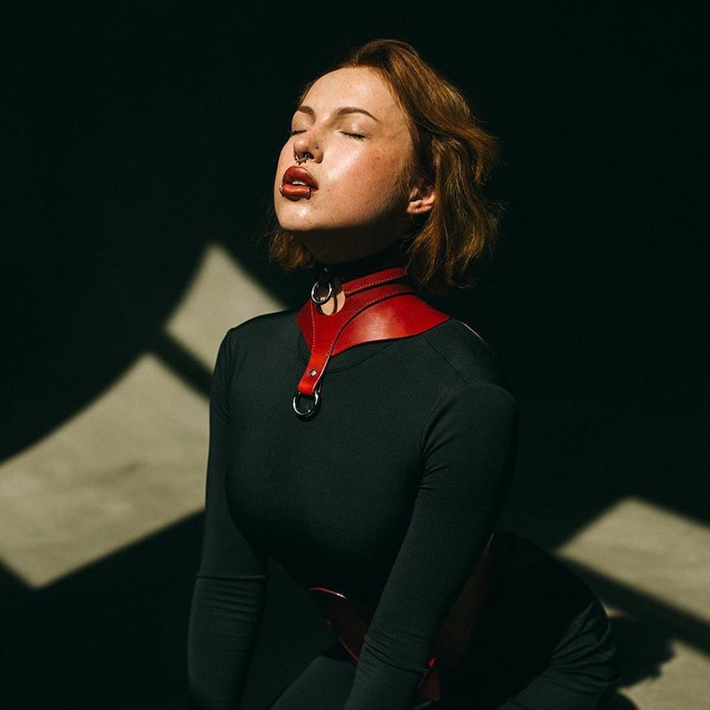 May include: A red leather collar featuring a wide, contoured neck band with a central vertical strap, silver-toned metal O-rings at the throat and chest, and a buckle closure, worn over a long-sleeved black top against a dark background.