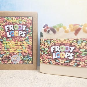 Peut inclure: Un pain de savon et sa boîte, tous deux avec le logo des céréales Froot Loops. Le savon est blanc avec des morceaux de céréales colorées sur le dessus. La boîte est marron avec le logo Froot Loops et des informations sur les ingrédients.