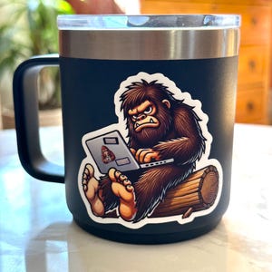 May include: A black travel mug with a sticker of a cartoon Bigfoot sitting on a log and using a laptop. The Bigfoot is brown and has a white beard and a grumpy expression.