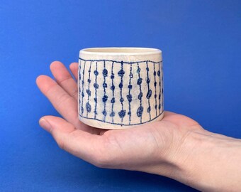 May include: A small, cylindrical ceramic cup with a cream-colored base and a blue patterned design. The design features vertical lines with small, irregular shapes. The cup is held in a hand against a blue background.