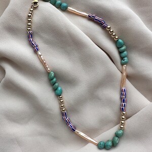 Op de afbeelding: Een handgemaakte ketting met turquoise, gouden en veelkleurige glaskralen. De ketting bevat ook delen van goudkleurige kralen en langwerpige, lichtgekleurde kralen. De ketting wordt gepresenteerd op een neutraal gekleurde stof.
