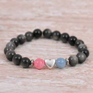 Puede incluir: Un brazalete de cuentas grises con una piedra rosa y azul y un dije de corazón plateado.