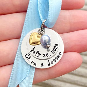 May include: A personalized wedding keepsake pendant with a light blue ribbon. The pendant features a gold heart, a pearl, and the names "Clara & Jesse" with the date "July 26, 2025" engraved on it. A charming wedding or anniversary gift.