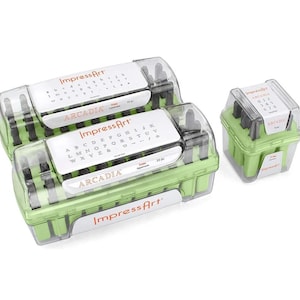 May include: Three light green plastic storage cases containing metal stamping tools. Two large rectangular cases hold uppercase and lowercase letters, while a smaller square case holds numbers. Each case features a clear lid with ImpressArt branding and a printed guide showing the Arcadia font characters included inside.