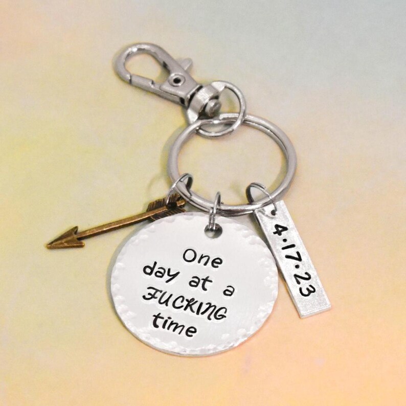 Peut inclure: Porte-cl&eacute;s argent&eacute; avec un charme rond qui dit "One day at a time" et un charme plus petit qui dit "4.17.23". Il y a aussi un petit charme en forme de fl&egrave;che attach&eacute; au porte-cl&eacute;s.