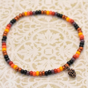 May include: A beaded bracelet featuring a mix of colorful beads in shades of orange, black, purple, and brown. A small, copper-colored pinecone charm hangs from the bracelet. The bracelet is displayed on a patterned surface.
