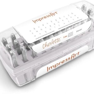 May include: A clear plastic storage case containing 33 metal stamps for jewelry making. The label reads ImpressArt, Charlotte, and 3 mm. The set includes lowercase letters and symbols, with a printed guide on the lid showing the font style.