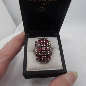 Puede incluir: Un anillo de plata con un grupo rectangular de piedras preciosas rojas rodeado de piedras preciosas negras más pequeñas. El anillo está en una caja negra con un interior blanco.